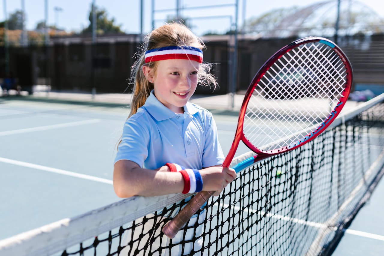 Smiling girl holding tennis racket by the net on a sunny court.
